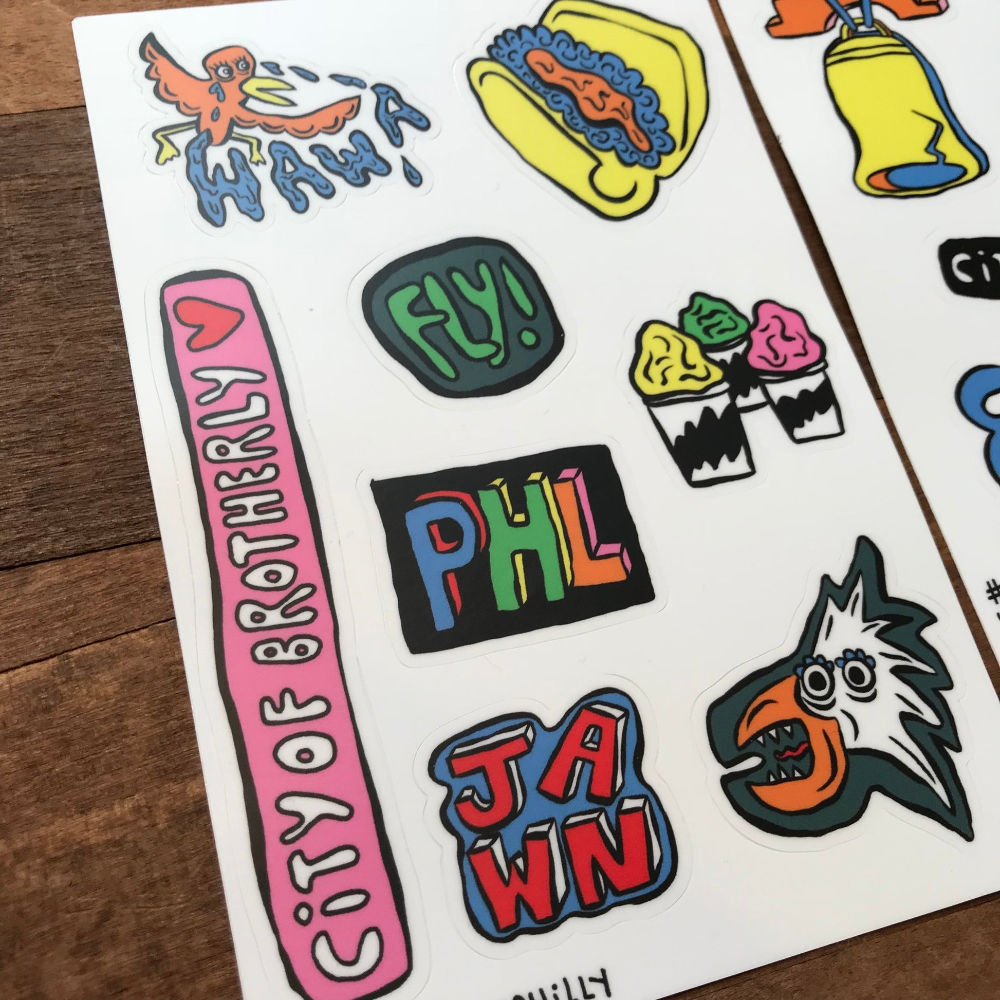 Assorted colorful Philly Sticker Packs with various designs and text, including "city of brotherly phl," "jawn," and a Philly-themed sticker pack featuring "Wawa" on a wood surface by Holly Simple.