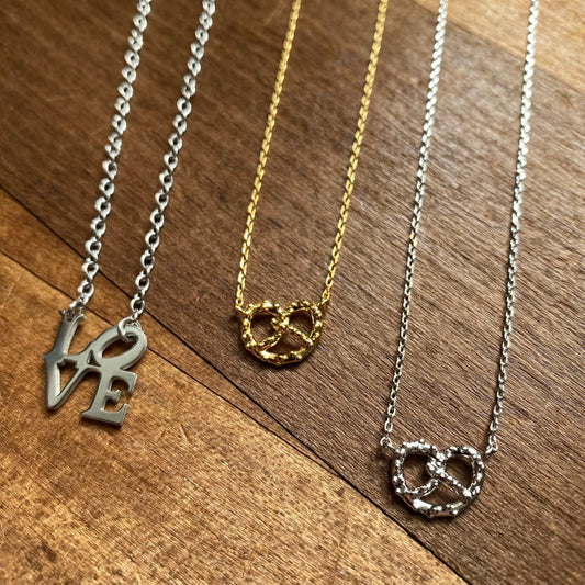 Three LOVE & pretzel necklaces with 'love' and infinity symbol designs on a wooden background, from Lily Lough Jewelry.