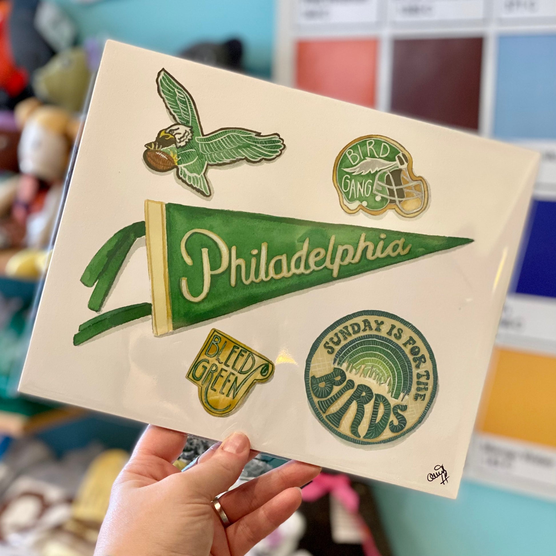 A hand holding a clear sheet adorned with colorful Meadowbrook Lane Philly Sports Prints, including logos and fan slogans on fine art paper.