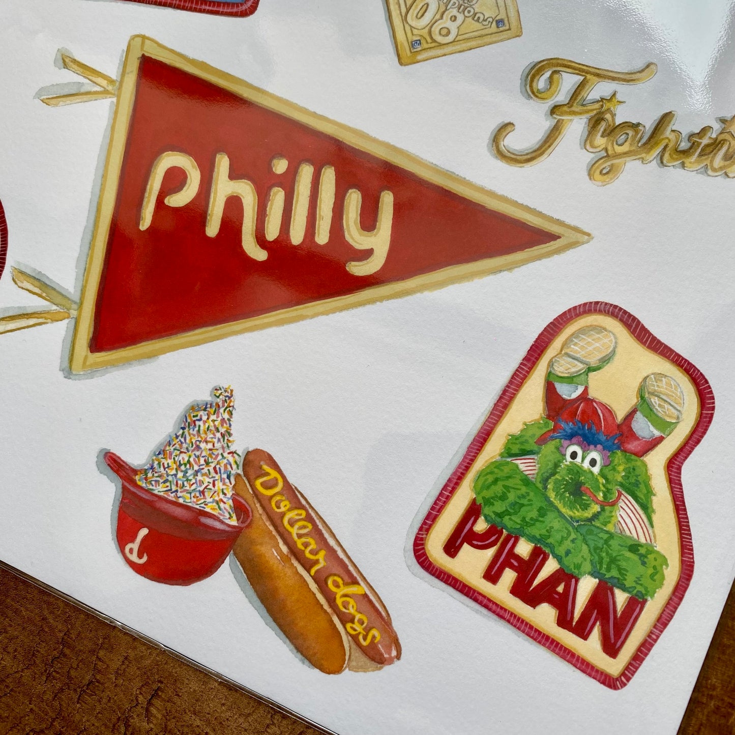 Illustrations featuring Philadelphia-themed items, including a pennant labeled 'Philly,' watercolor prints of a water ice, a hot dog, and a character wearing a Philadelphia Phillies jersey, all meticulously rendered by Meadowbrook Lane's Philly Sports Prints.