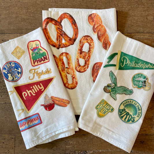 Two Philly Illustrated Tea Towels from Meadowbrook Lane displaying Philadelphia-themed designs, one with pretzels and the other featuring sports team logos and local icons.