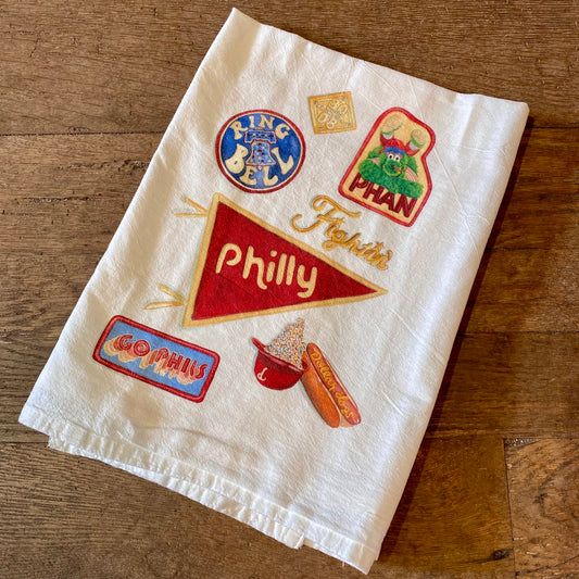 A folded Philly Illustrated Tea Towel with embroidered patches featuring Philadelphia-themed icons and text from Meadowbrook Lane.