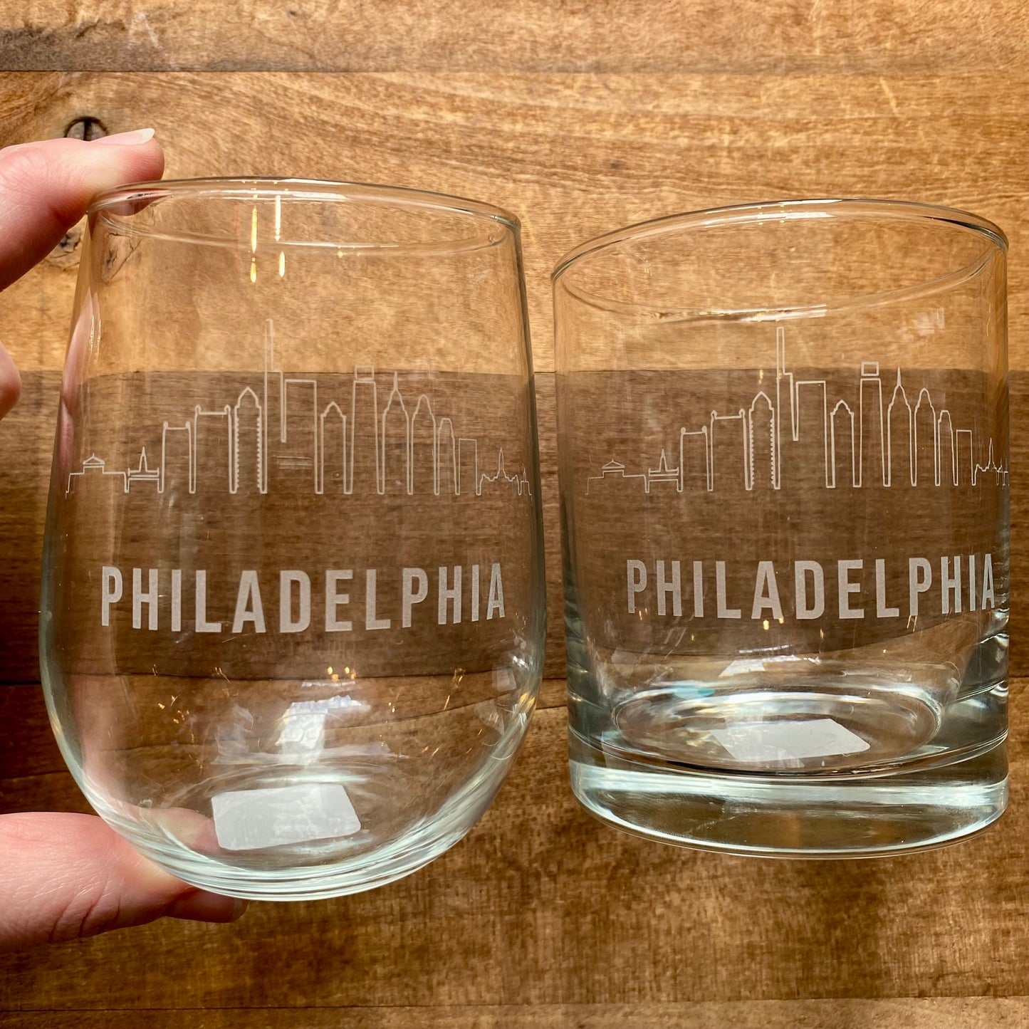 Two Philly Etched Wine & Rocks Glasses from Morning Fog Studios, featuring a skyline design, with one being held and the other on a wooden surface.