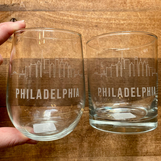 Two Philly Etched Wine & Rocks Glasses from Morning Fog Studios, featuring a skyline design, with one being held and the other on a wooden surface.