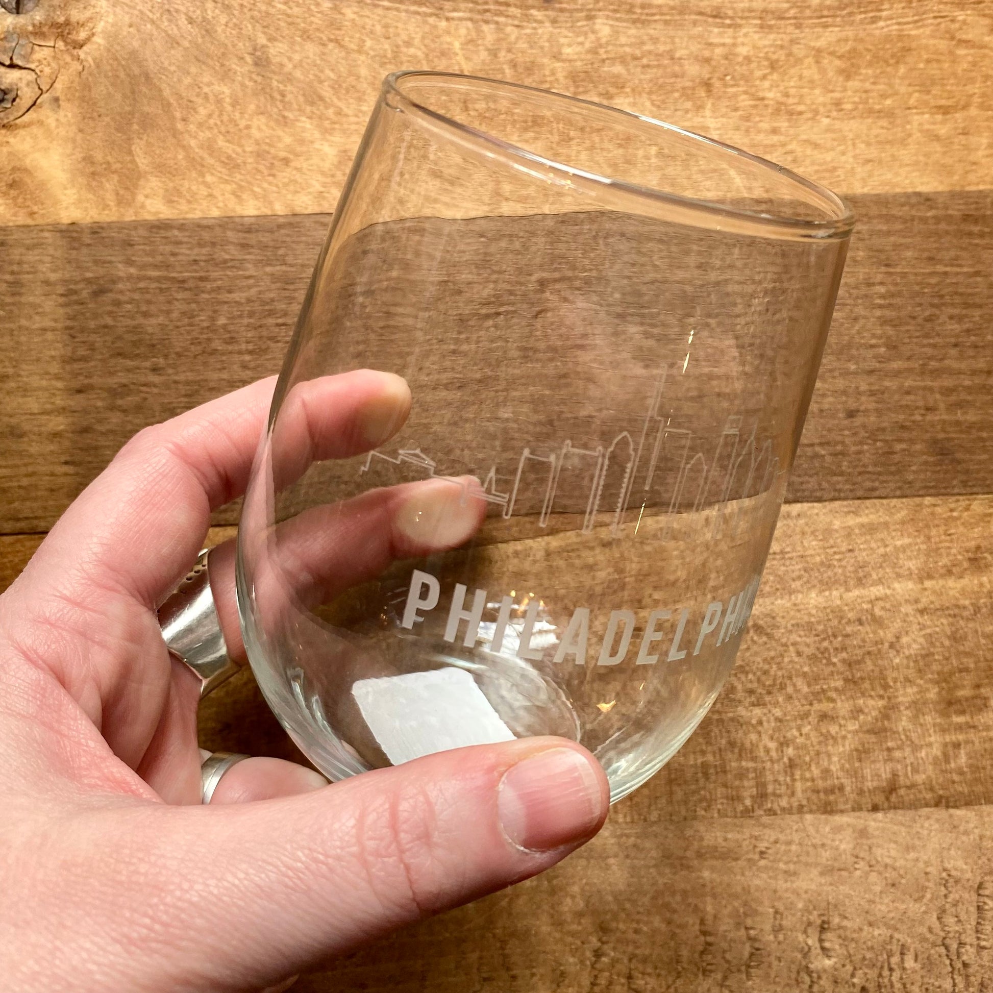 A hand holding a Philly Etched Wine & Rocks Glassware with the "Philadelphia skyline" etched on it, placed on a wooden surface, made by Morning Fog Studios.