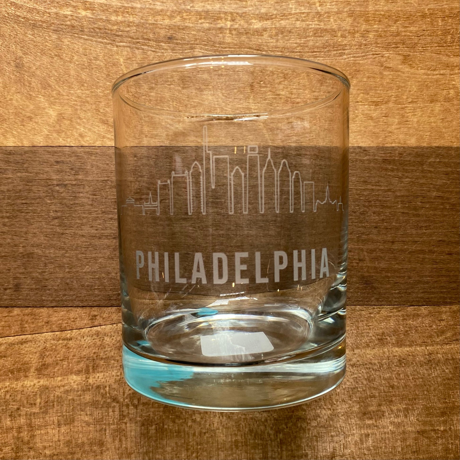 A Philly Etched Wine & Rocks Glassware with the Philadelphia skyline, from Morning Fog Studios, placed on a wooden surface.