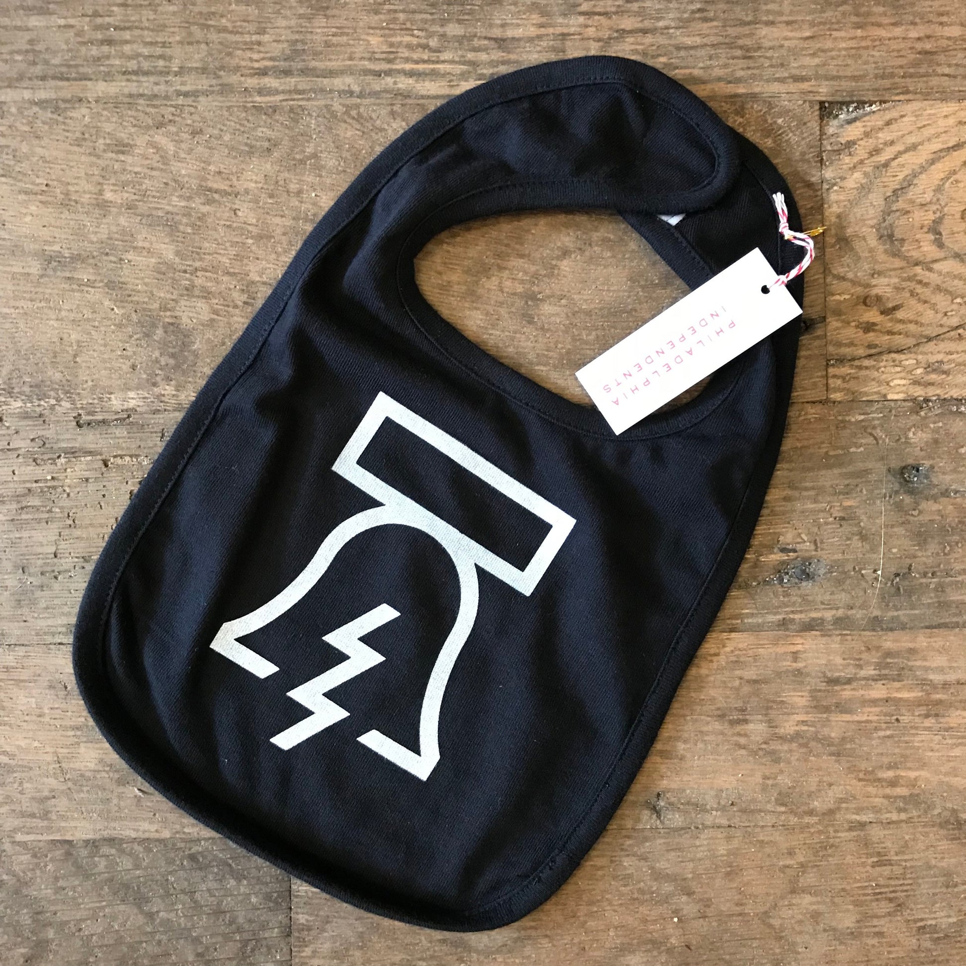 A Bell & Bolt Baby Bib in black cotton with a white outline of a superhero cape design and a tag attached to it by Philadelphia Independents.