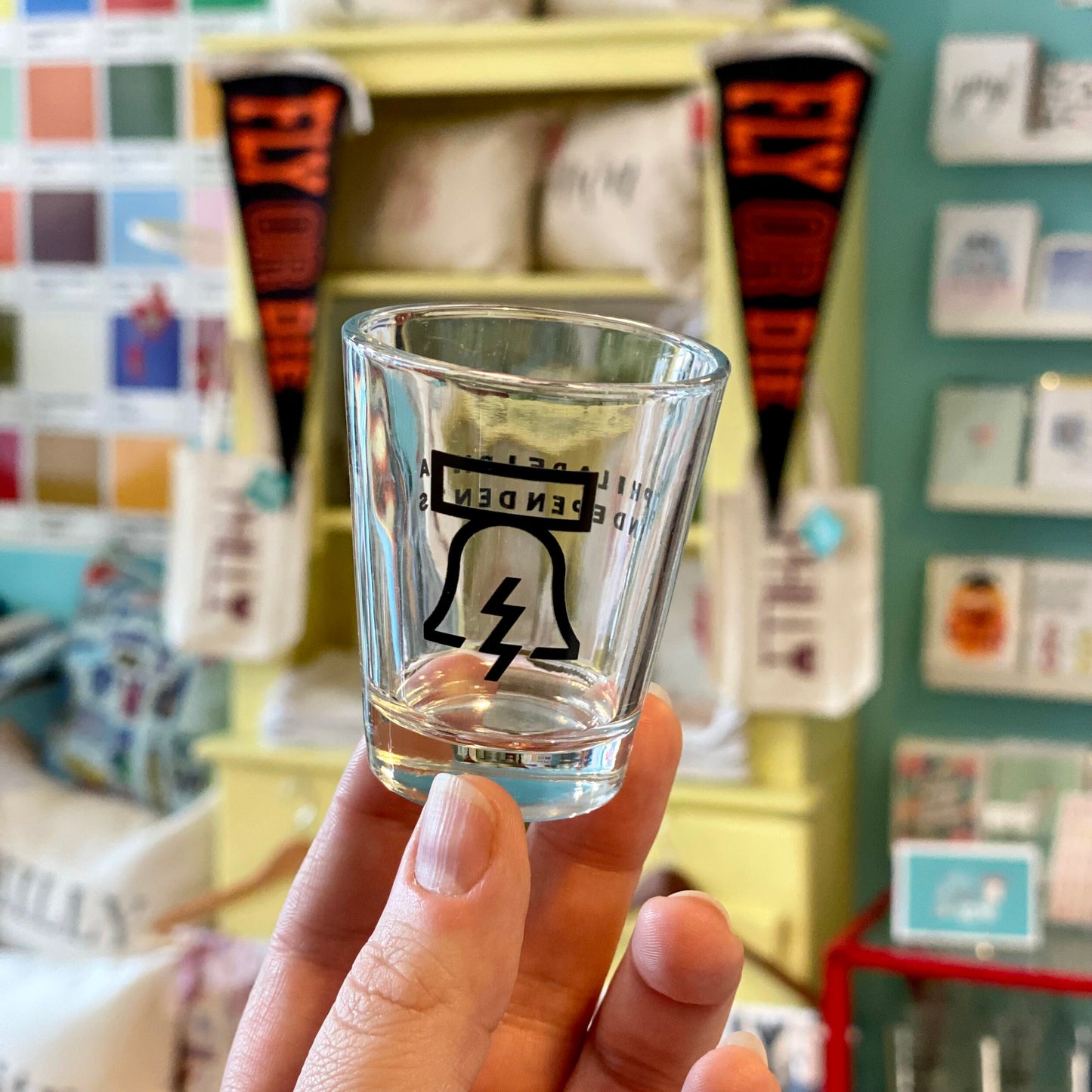 A dishwasher-safe Bell & Bolt Shot Glass with a lightning bolt design being held in a colorful Philadelphia Independents retail setting.
