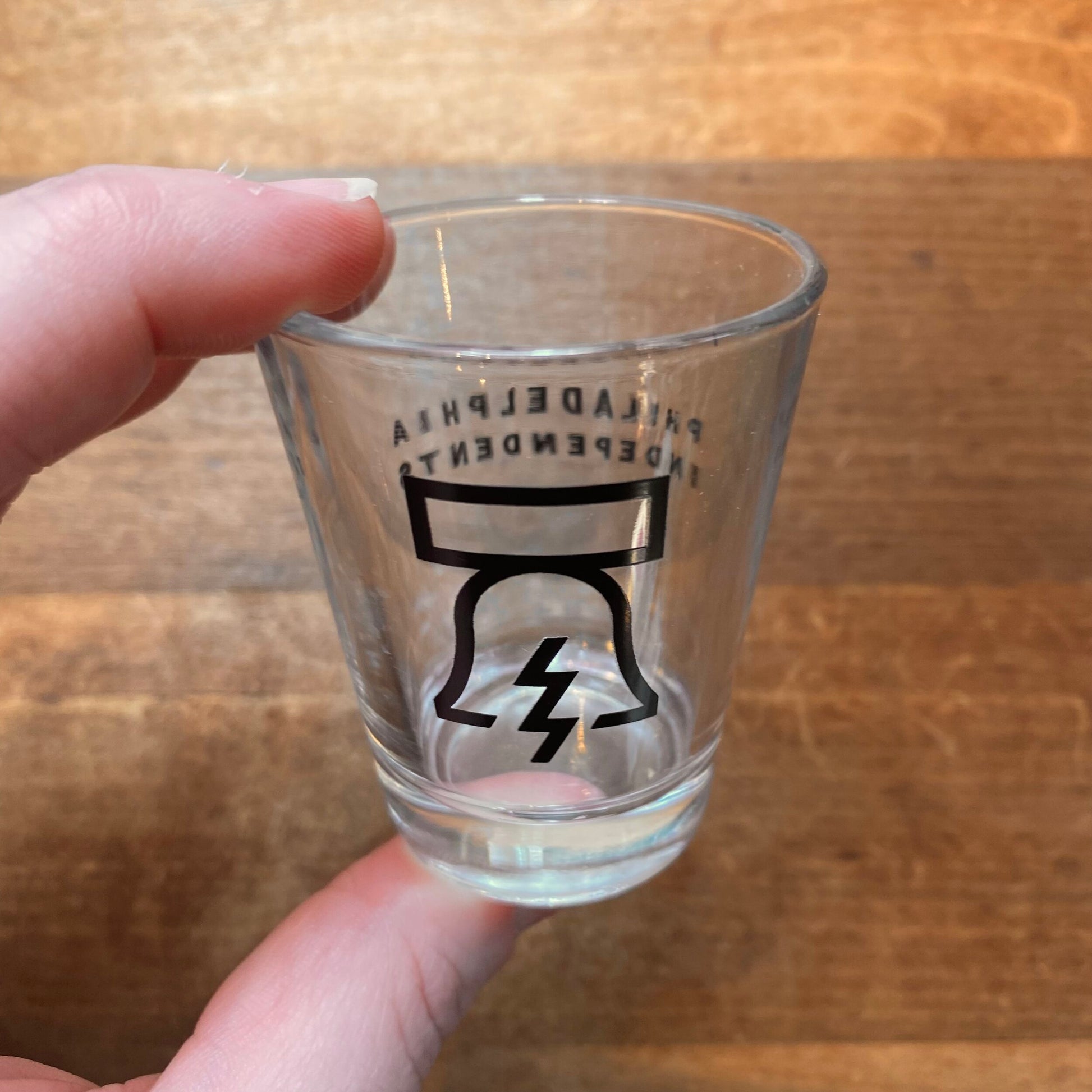 A hand holding a Philadelphia Independents Bell & Bolt Shot Glass with a lightning bolt design and stylized text above it.