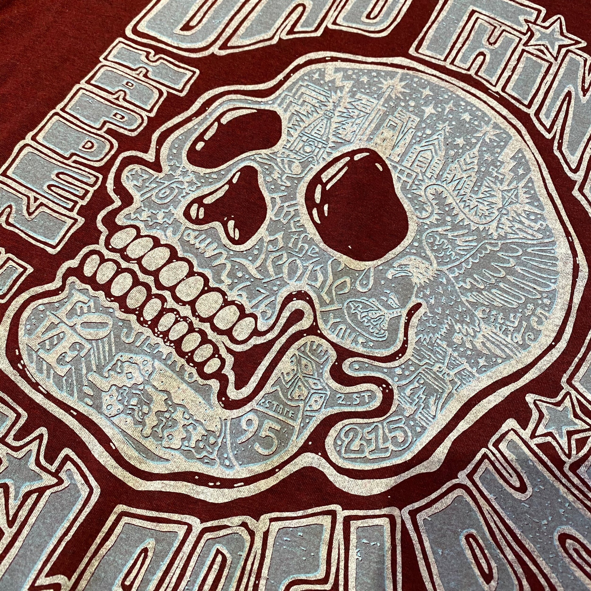 A printed graphic of a stylized skull with elaborate patterns on a Paul Carpenter Bad Things Happen T-Shirt made of Bella & Canvas Tri-Blend maroon fabric.