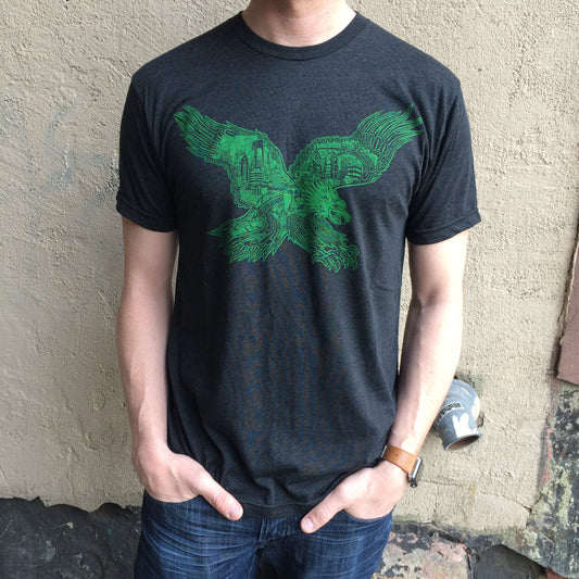 Man wearing a Paul Carpenter Swooping Eagle T-Shirt in charcoal gray with a green graphic print of a Philadelphia eagle in front of a building facade.
