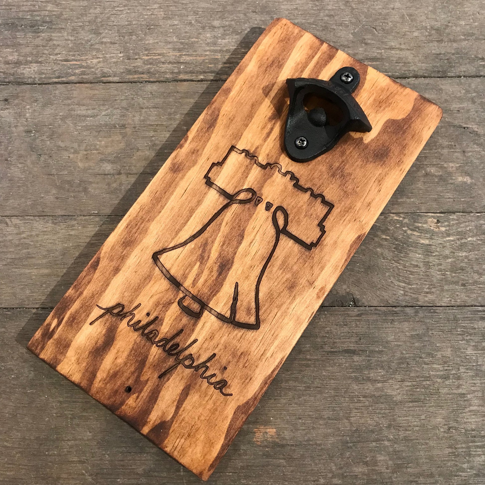 A Philly Wall-Mounted Bottle Opener with a laser-etched image of the Liberty Bell and "Philadelphia" text, mounted on a rustic wood plank, from Philly Phlights.