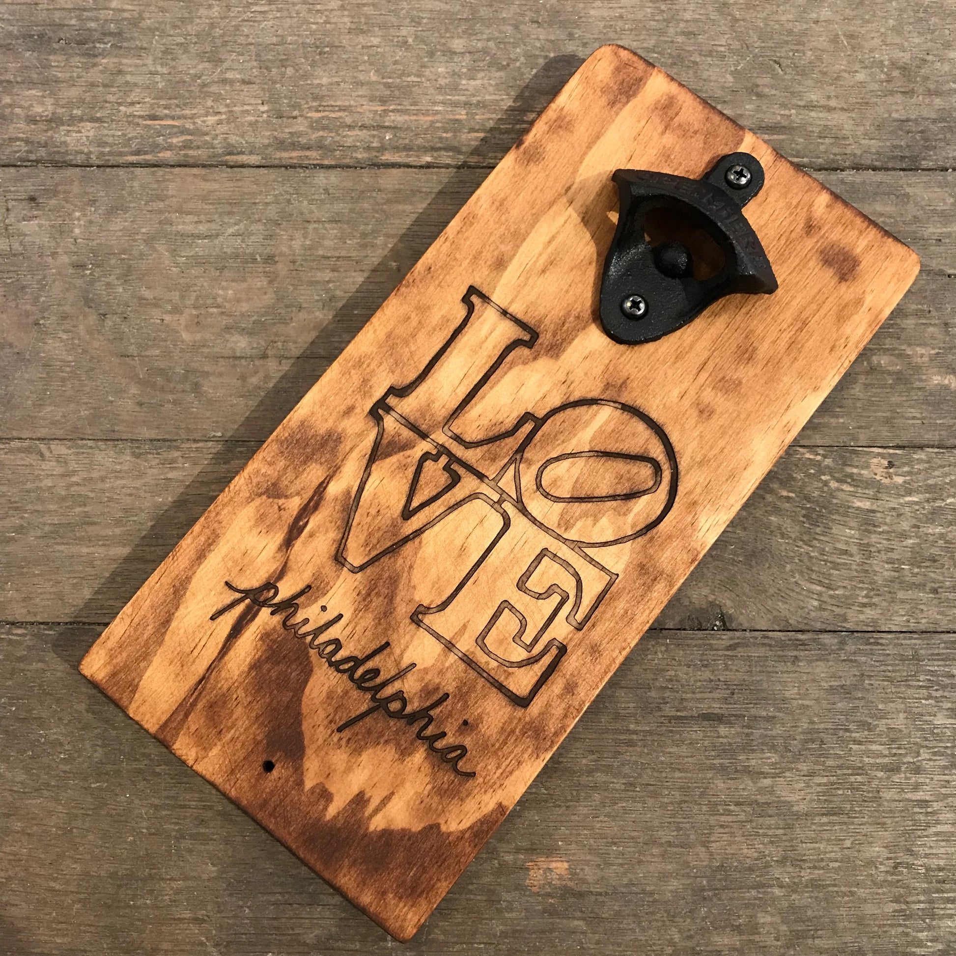 A Philly Phlights Philly Wall-Mounted Bottle Opener with a "love" design and the inscription "friendship" laser-etched into the surface, resting on a wooden floor.