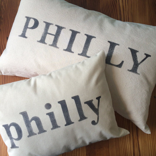 Two light-colored pillows rest on a wooden surface. The larger pillow, made from unbleached cotton, showcases "PHILLY" in uppercase letters, while the smaller Philly pillow displays "philly" in lowercase letters. Both are crafted by The Pillow Works for a charming and cohesive look.