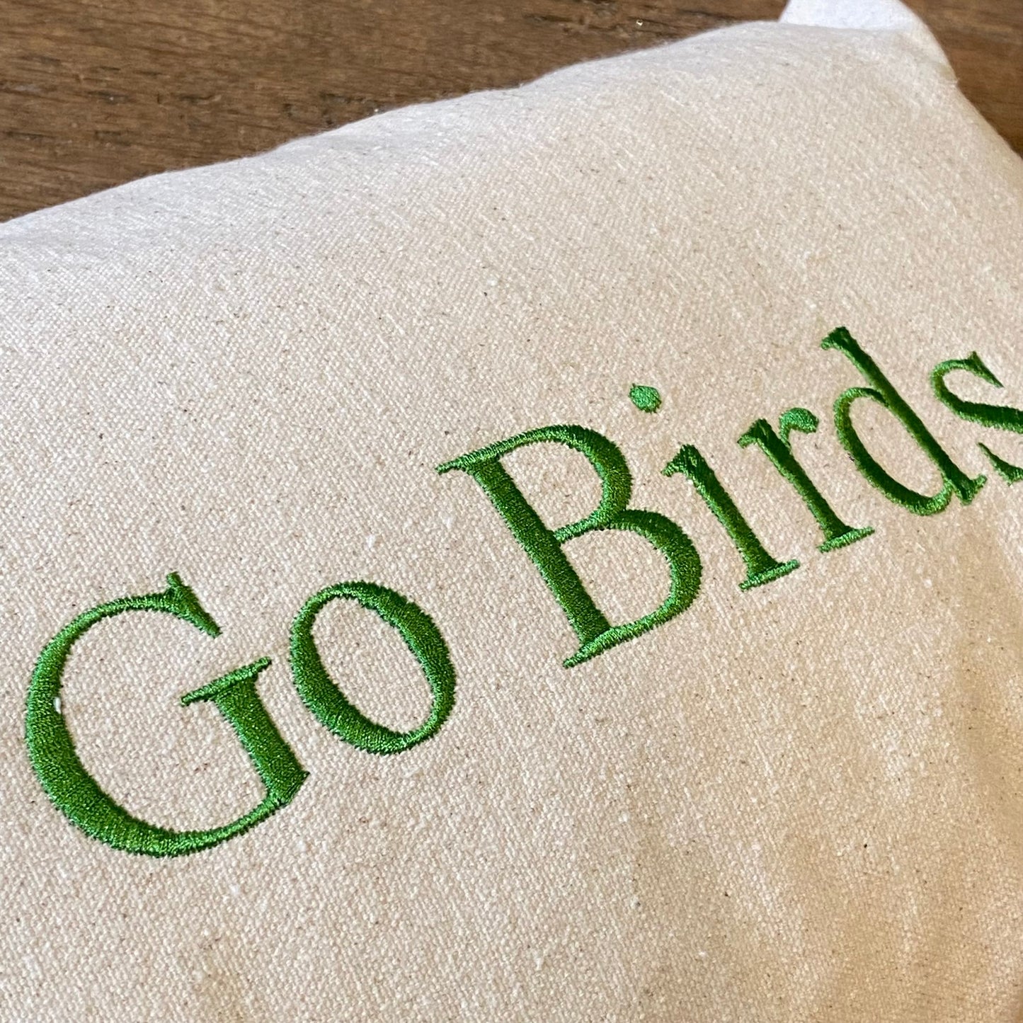 A close-up of a beige 100% unbleached cotton drill fabric with the phrase "Go Birds" embroidered in kelly green thread on The Pillow Works Go Birds Embroidered Pillow.