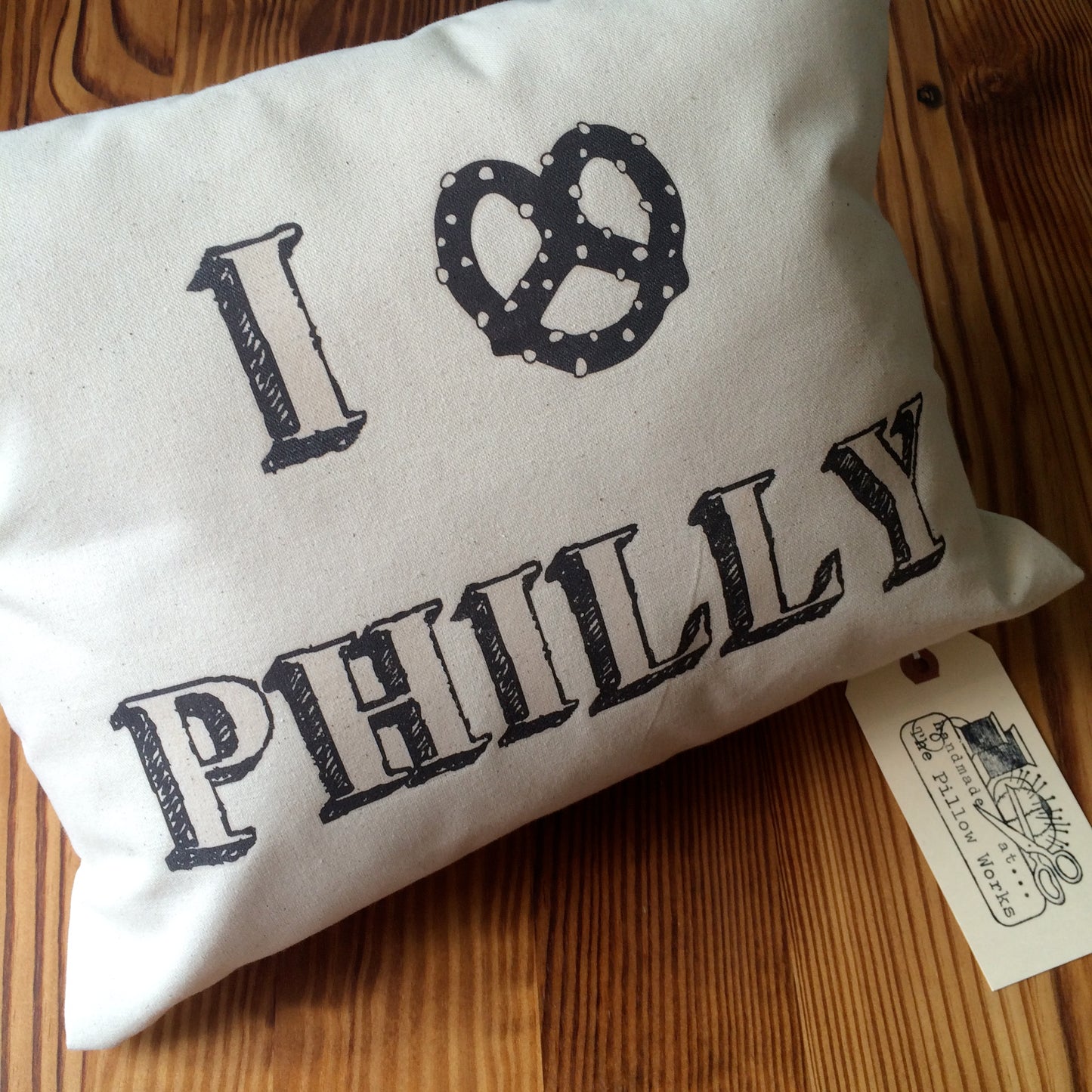 White I Pretzel Philly Pillow with "I Pretzel Philly" printed in black, placed on a wooden surface, with a brown tag attached. Made by The Pillow Works.