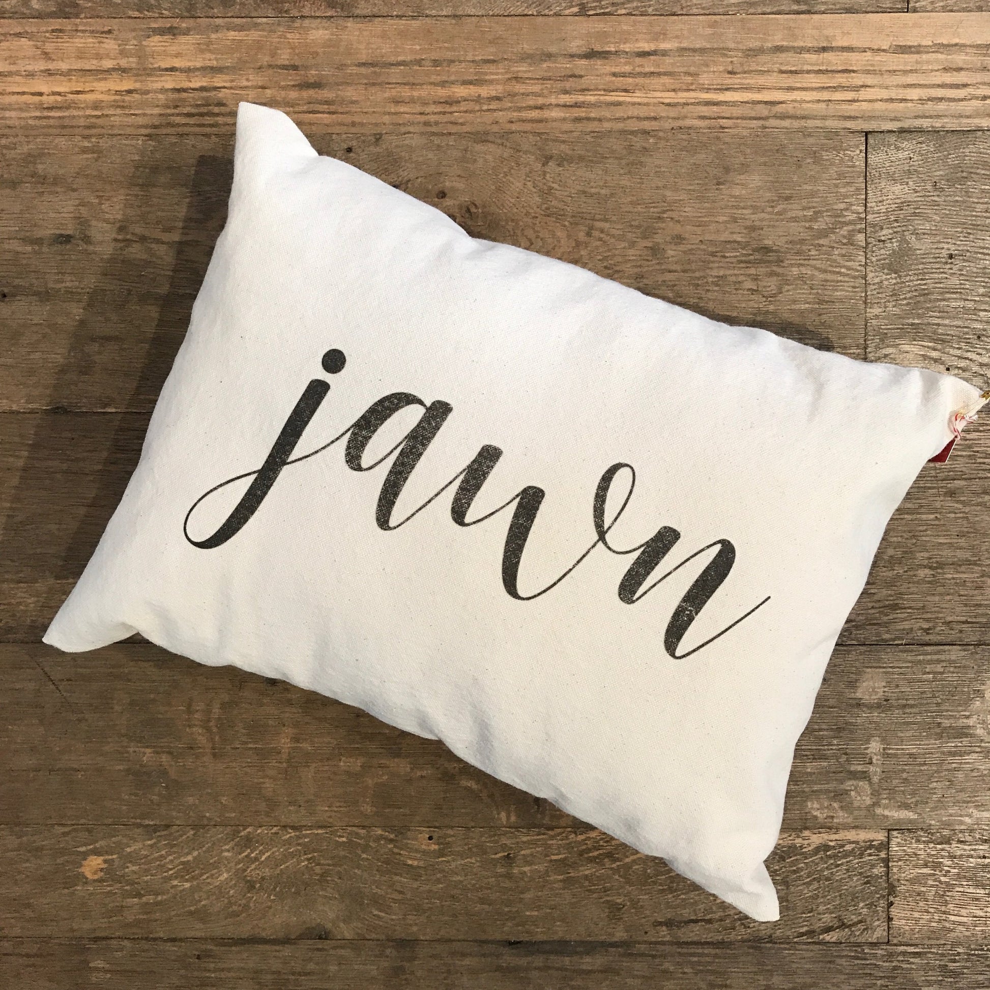 A Jawn Pillow by The Pillow Works, a rectangular white accent pillow made from cotton drill fabric and adorned with the word "jawn" written in elegant cursive, rests gracefully on a wooden floor.