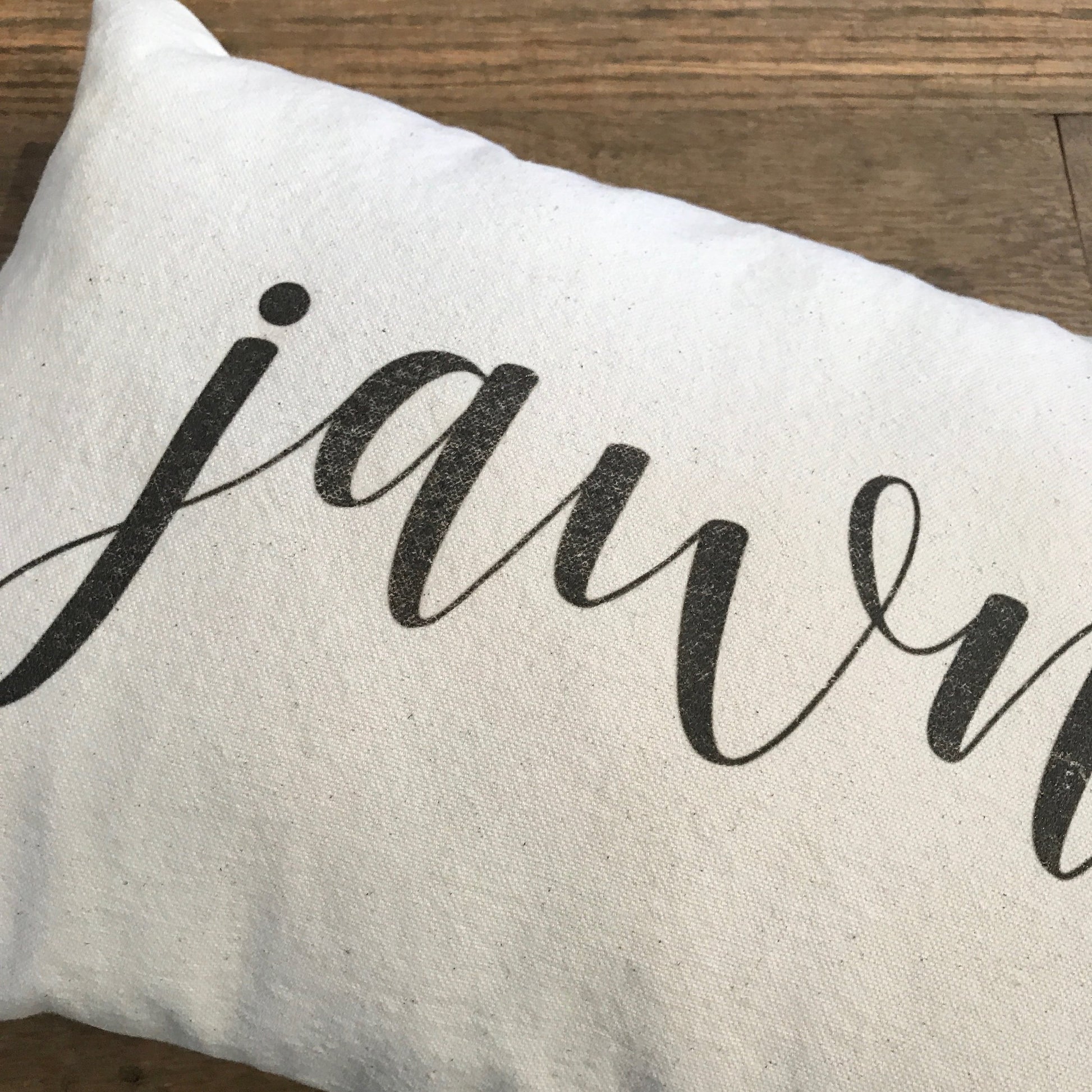 A **Jawn Pillow** with the word "jawn" elegantly written in black cursive letters lies on a wooden surface, crafted from soft cotton drill fabric by **The Pillow Works**.