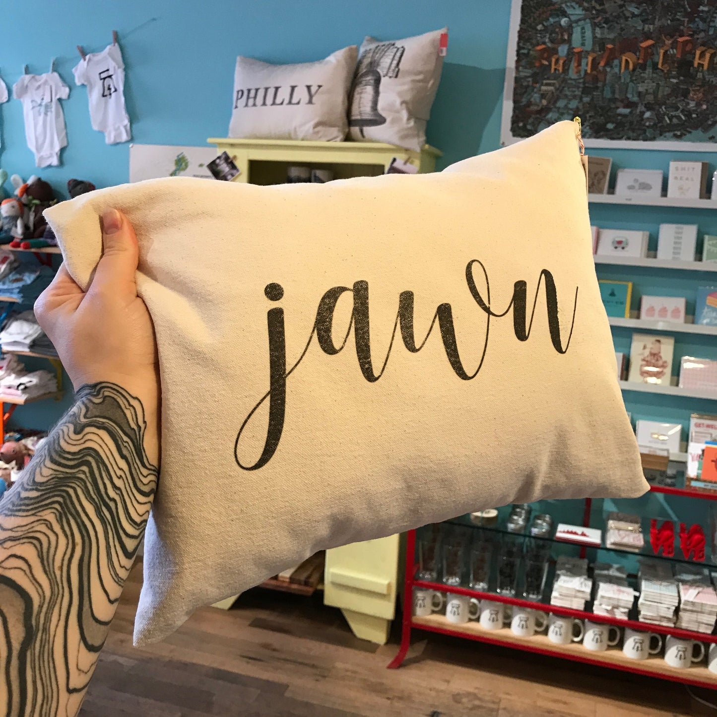 A tattooed hand holds a white Jawn Pillow by The Pillow Works with the word "jawn" elegantly written in cursive, set against a background of various items on shelves in a store.