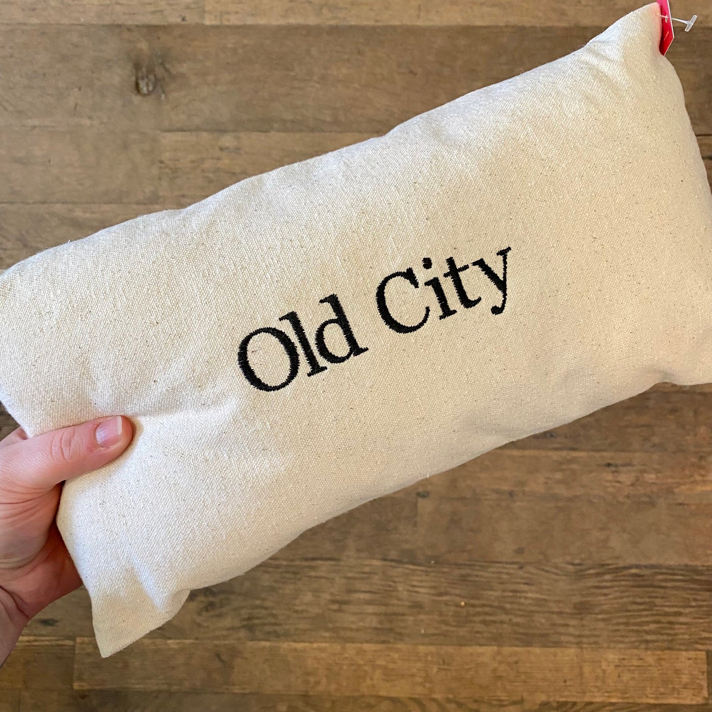 A hand holding a beige embroidered Old City pillow from The Pillow Works with "Old City" printed in black text.