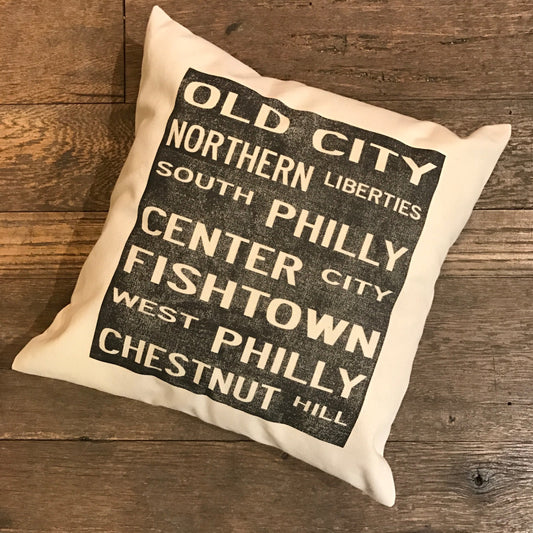 Square Philadelphia Neighborhoods Pillow featuring black text with Philly neighborhoods listed on unbleached cotton drill fabric, set on a wood floor by The Pillow Works.