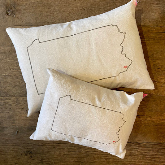 Two Pennsylvania Heart Pillows from The Pillow Works on a wooden surface, one featuring an embroidered outline of Pennsylvania with a small red heart detail, made from unbleached cotton.