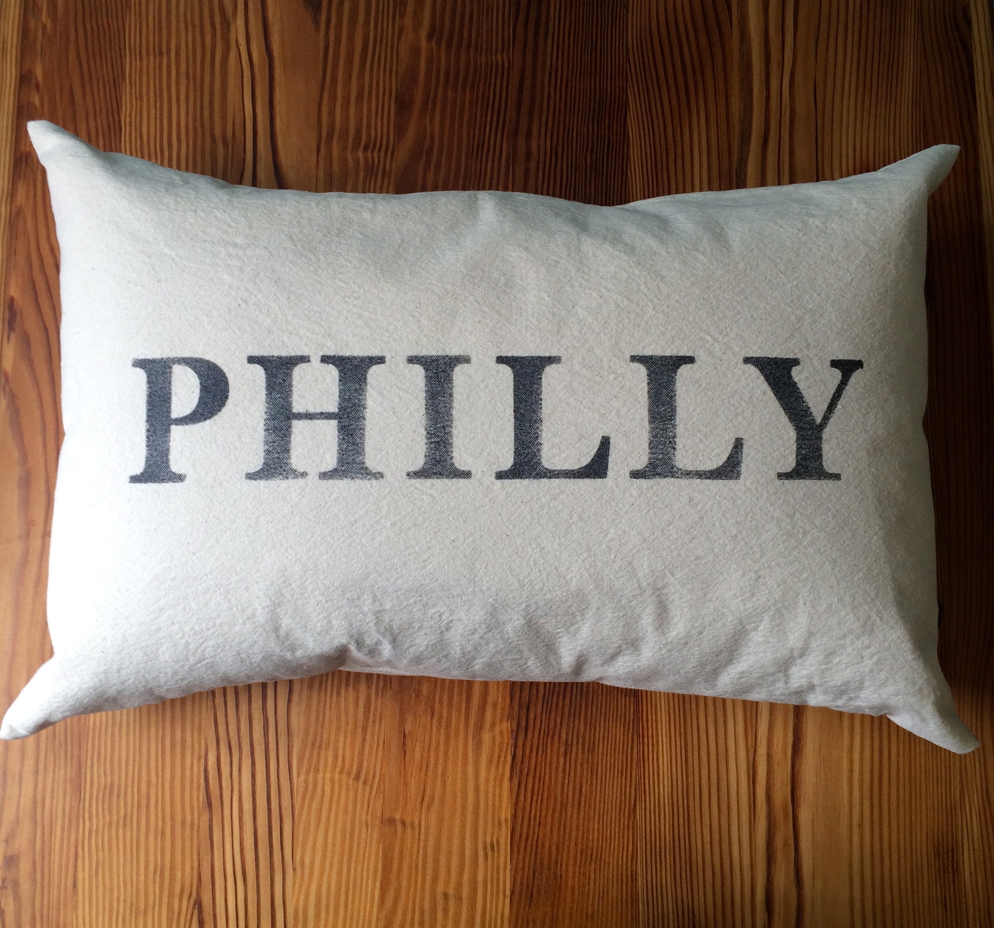 Rectangular unbleached cotton Philly Pillows with the word "PHILLY" printed in bold, black letters, set against a wooden background from The Pillow Works.