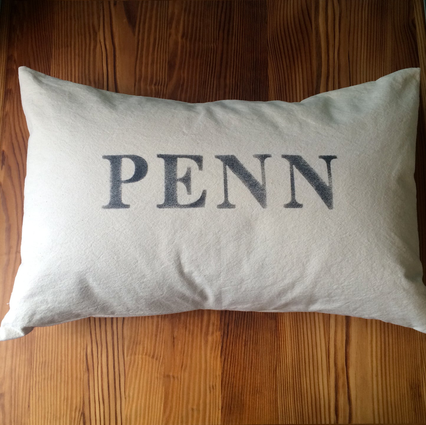 A rectangular pillow with the word "Penn" printed in dark letters, celebrating school pride, resting on a wooden surface from The Pillow Works School Pride Pillows.