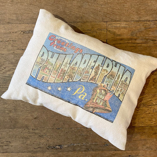 A Philly Postcard Pillow from The Pillow Works, featuring a vintage "greetings from Philadelphia" postcard design on unbleached cotton, placed on a wooden floor.