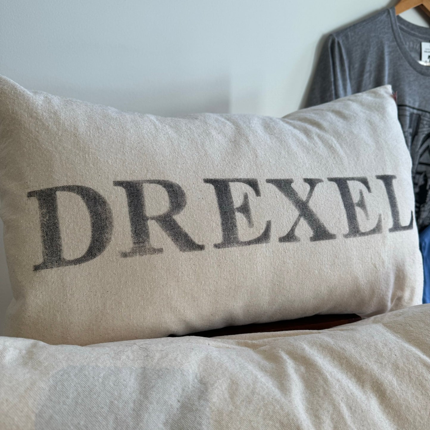 A close-up of a rectangular The Pillow Works School Pride Pillow with the word "Drexel" printed in gray, placed on a wooden surface against a blurred background.