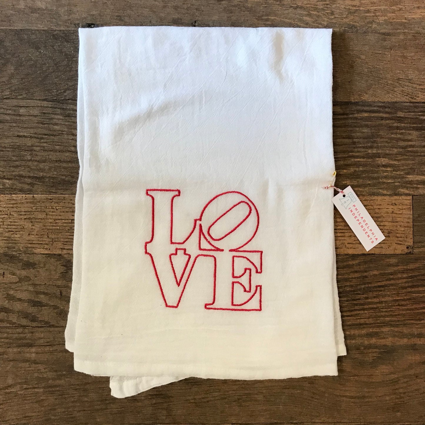 A white 100% cotton flour sack LOVE Tea Towel from The Pillow Works embroidered in red on a wooden surface with a price tag attached.