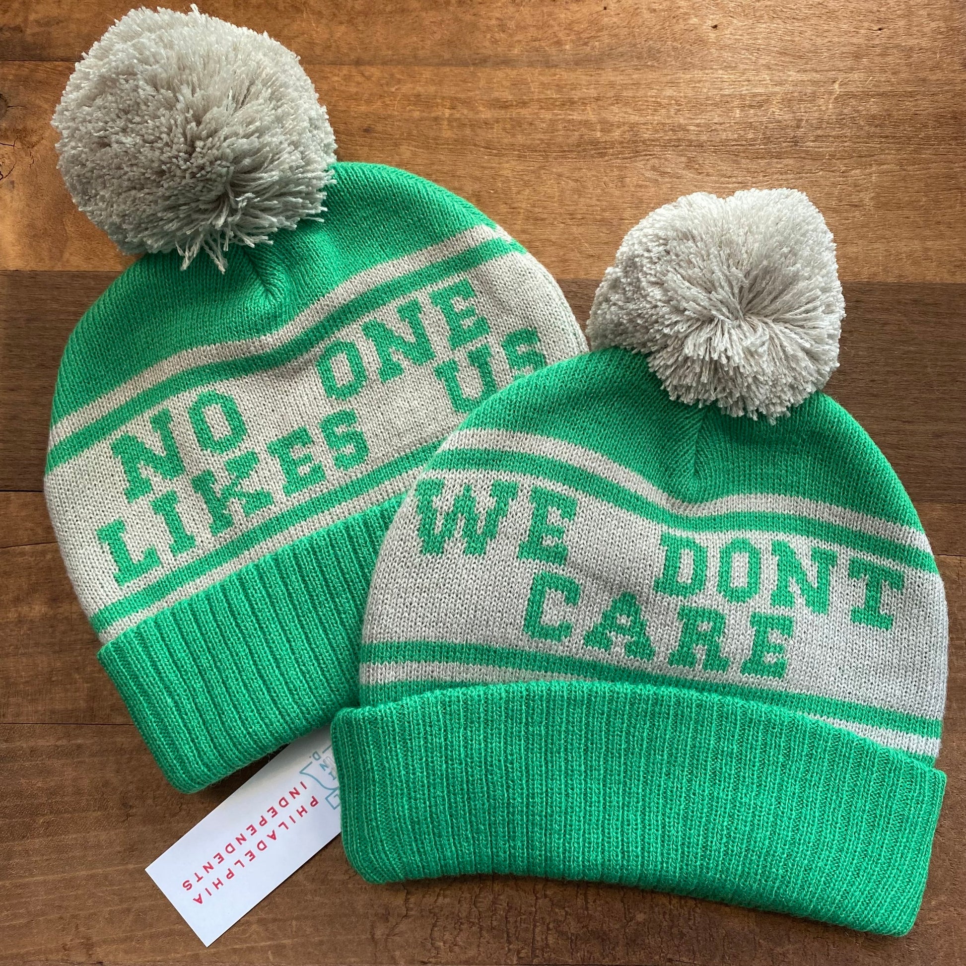 Two green and white South Fellini Philadelphia Eagles Baby No One Likes Us Beanie hats with pompoms, one reads "no one likes us" and the other "we don't care", on a wooden surface.