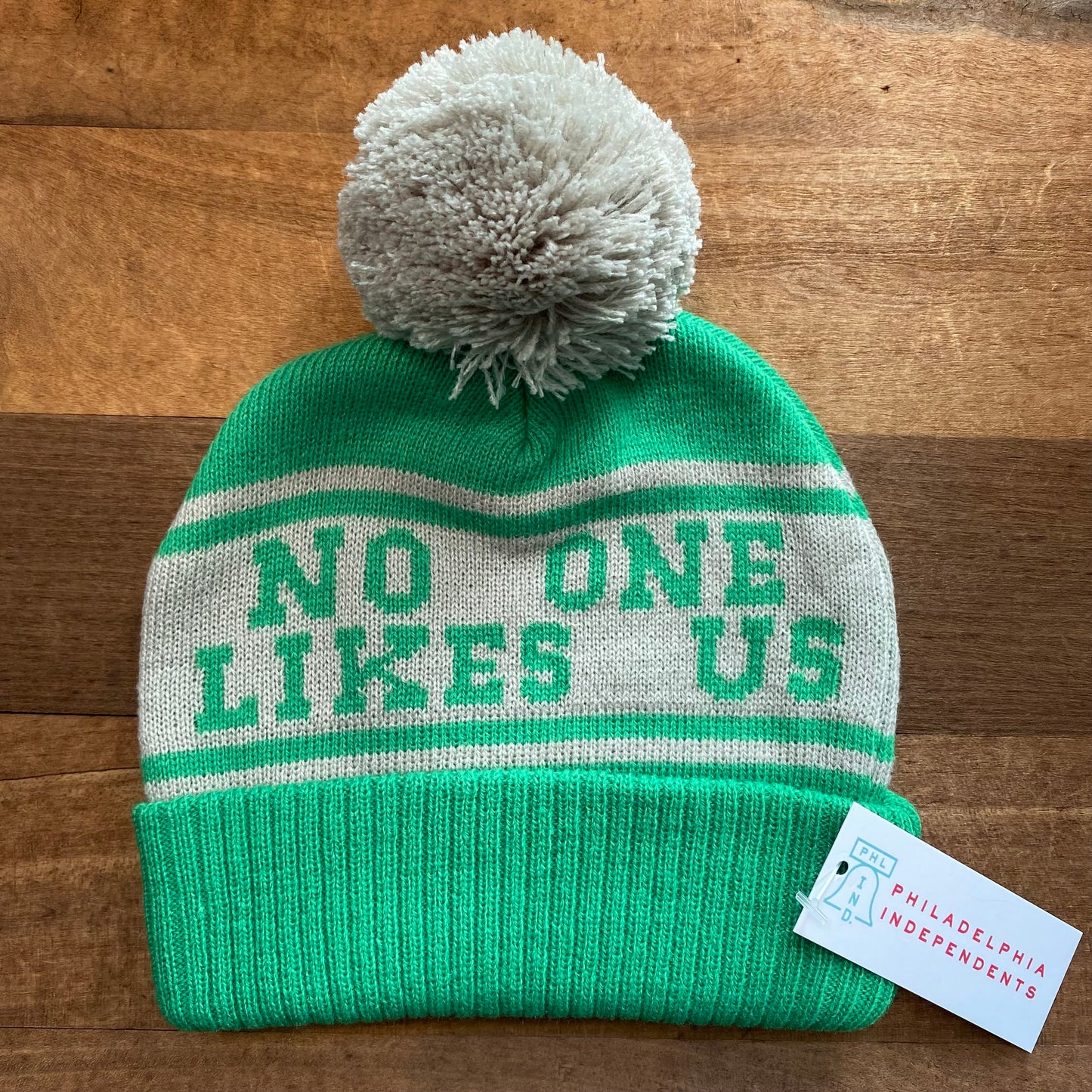 A green and white knit hat with a pom-pom and the phrase "no one likes us," reminiscent of Jason Kelce's famous sentiment, displayed, resting on a wooden surface. (Product Name: Baby No One Likes Us Eagles Beanie by South Fellini)