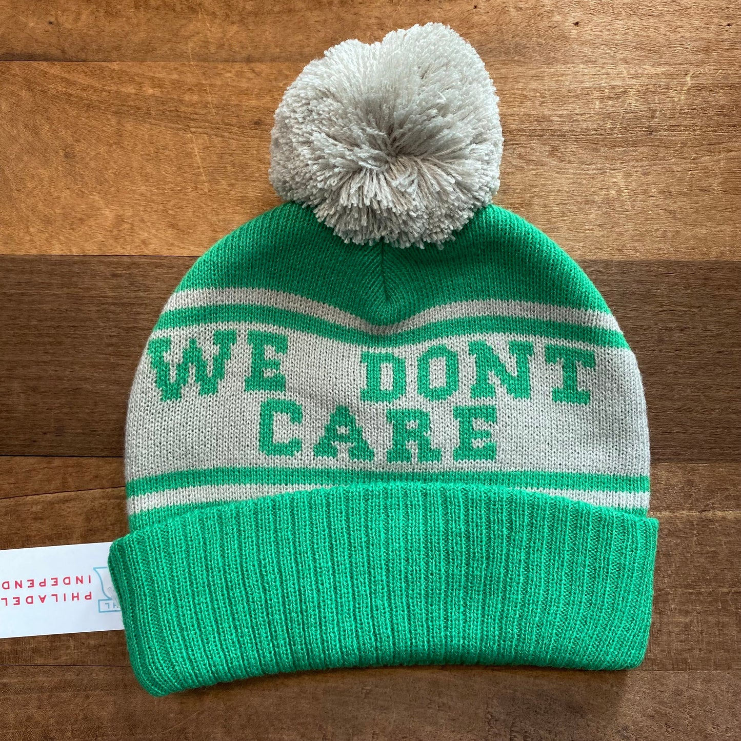 Baby No One Likes Us Eagles Beanie by South Fellini, symbolizing the Philadelphia Eagles colors, with a pompom and the phrase "we don't care" woven into the design, displayed on a wooden surface.