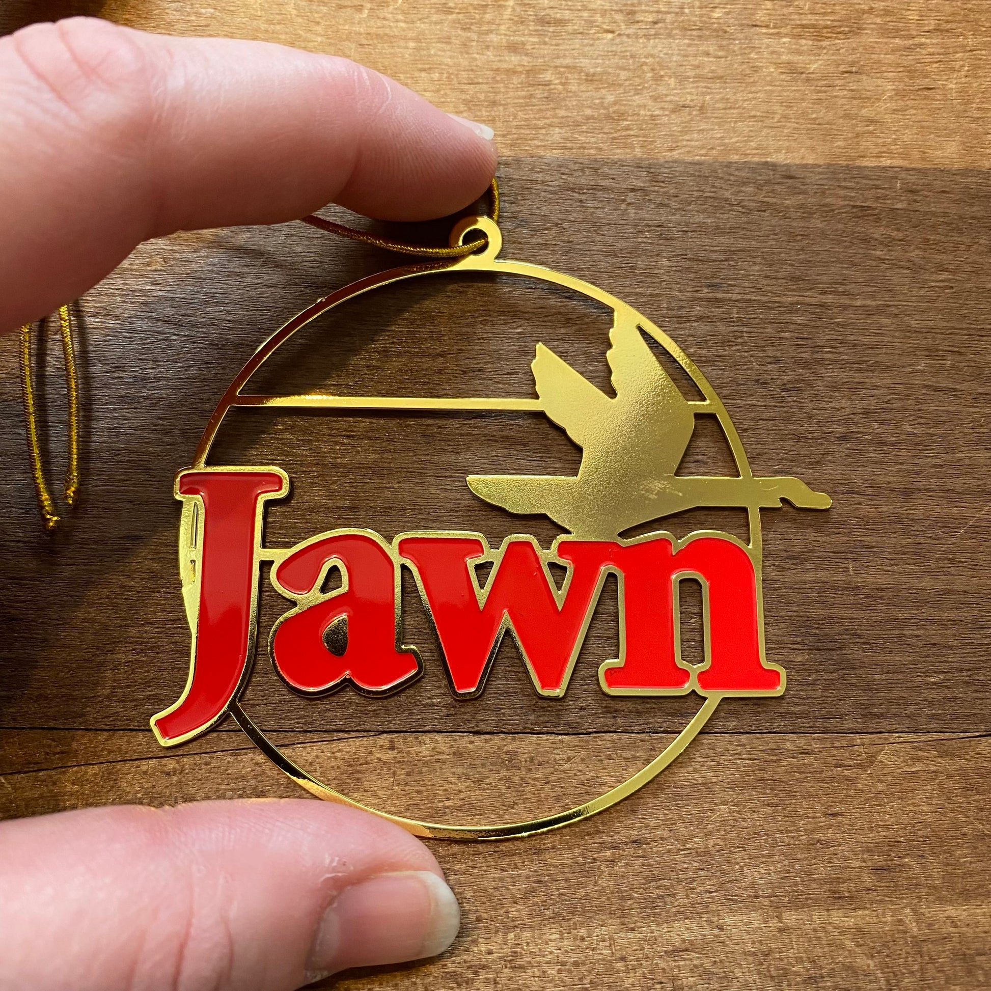Gold and red South Fellini Jawn Ornament reading "jawn" with a silhouette of a plane, held by a person against a wooden background.