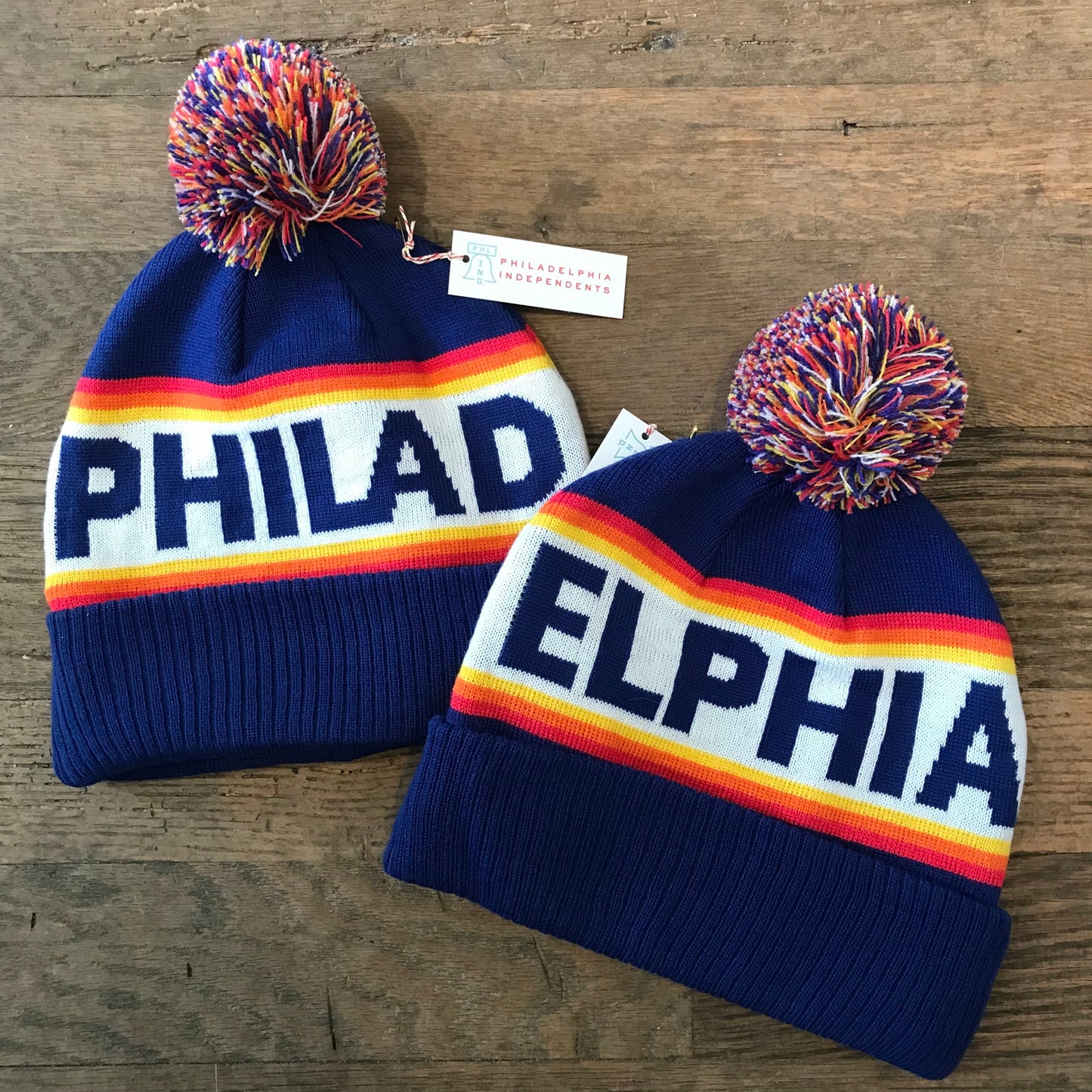 Two blue, double-sided knit South Fellini winter hats with pom-poms and "Philadelphia" written across them, displayed on a wooden surface.