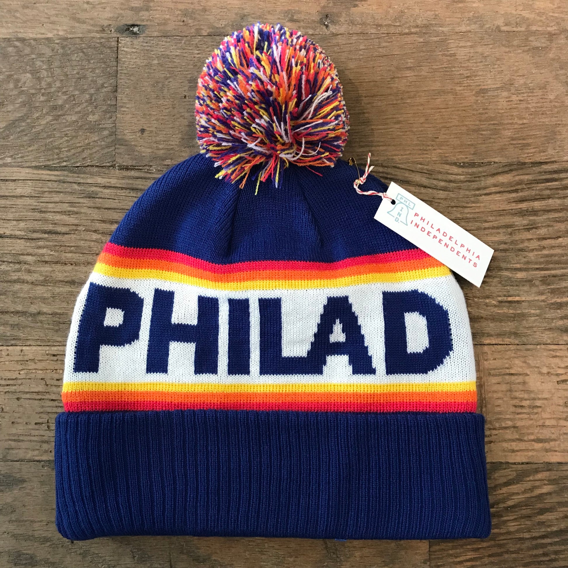 New blue double-sided knit Philadelphia Beanie with colorful pom-pom and "South Fellini" text lying on a wooden surface.