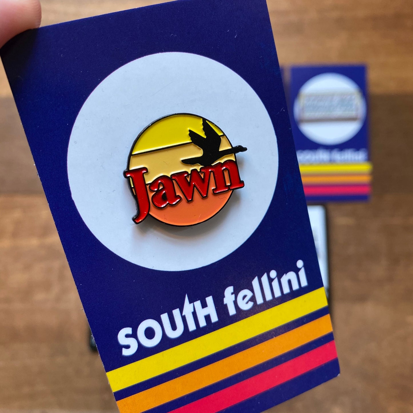 A hand holds a South Fellini Philly-Themed Enamel Pin with the word "jawn" inside a circular design, above the text "South Fellini" and colorful stripes. Other stickers are partially visible in the background.