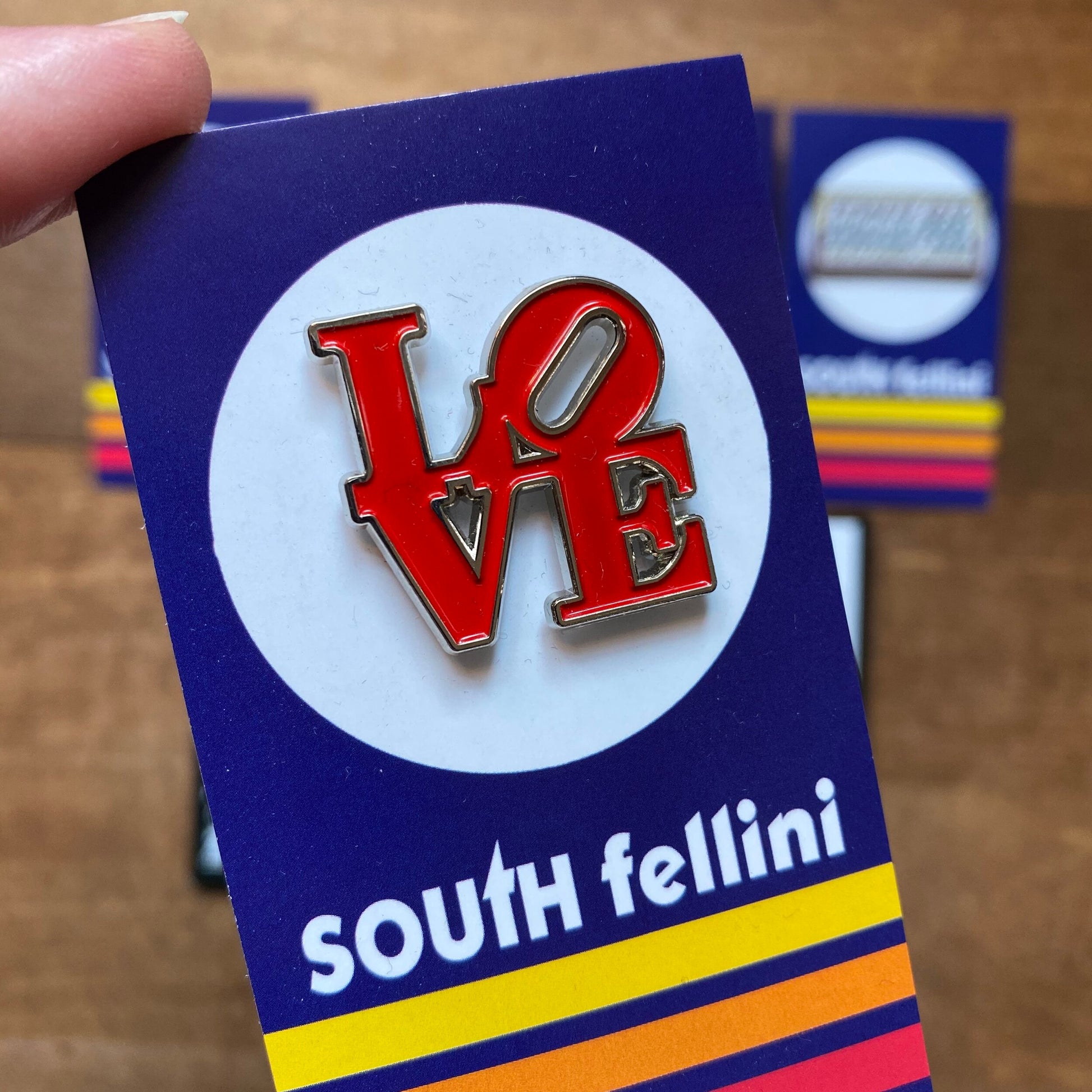 A hand holding a card with a "love" Philly-Themed Enamel Pin attached, featuring the brand "South Fellini" below multicolored stripes.