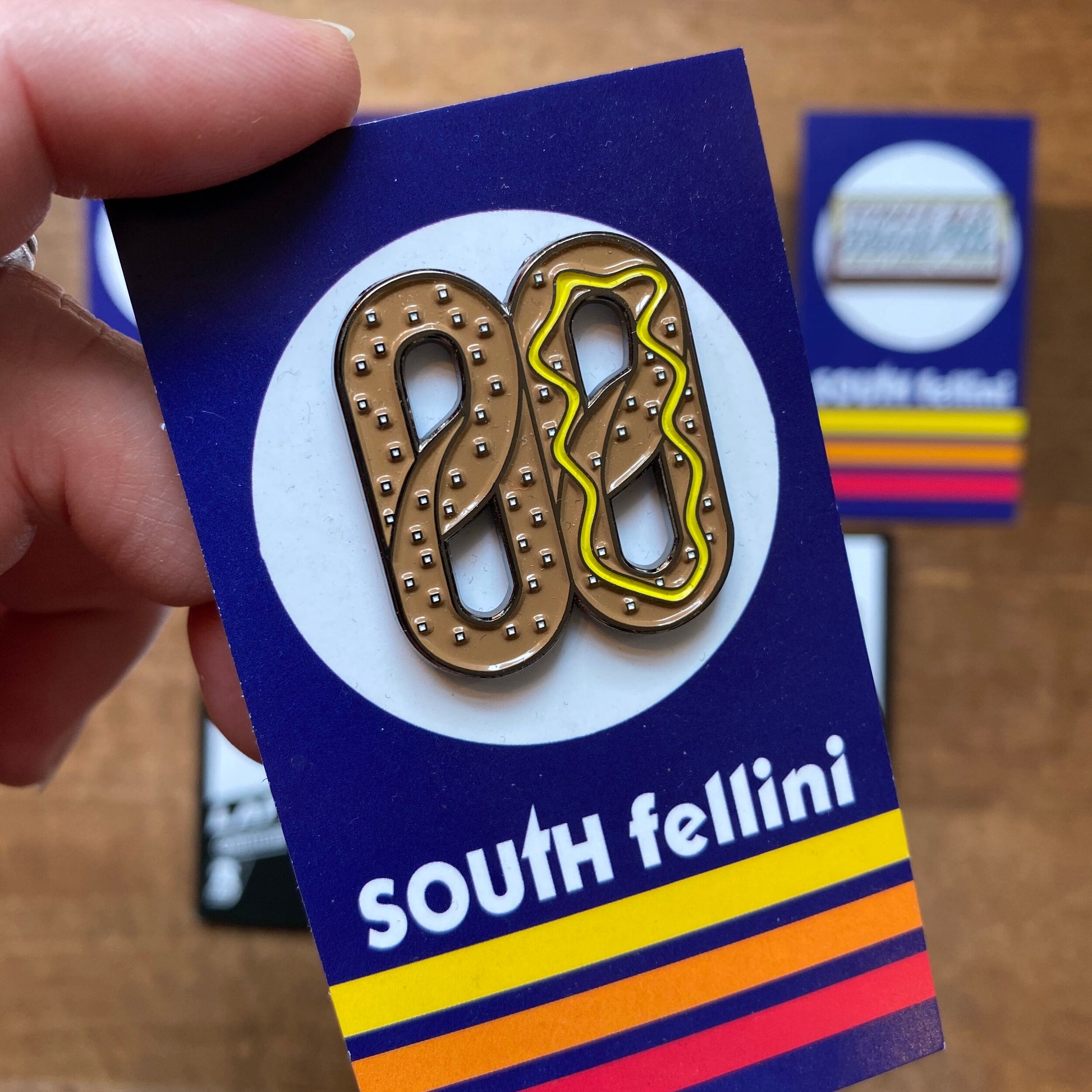 Hand holding a South Fellini Philly-Themed Enamel Pin with a pretzel-shaped "88" design on a colorful background.