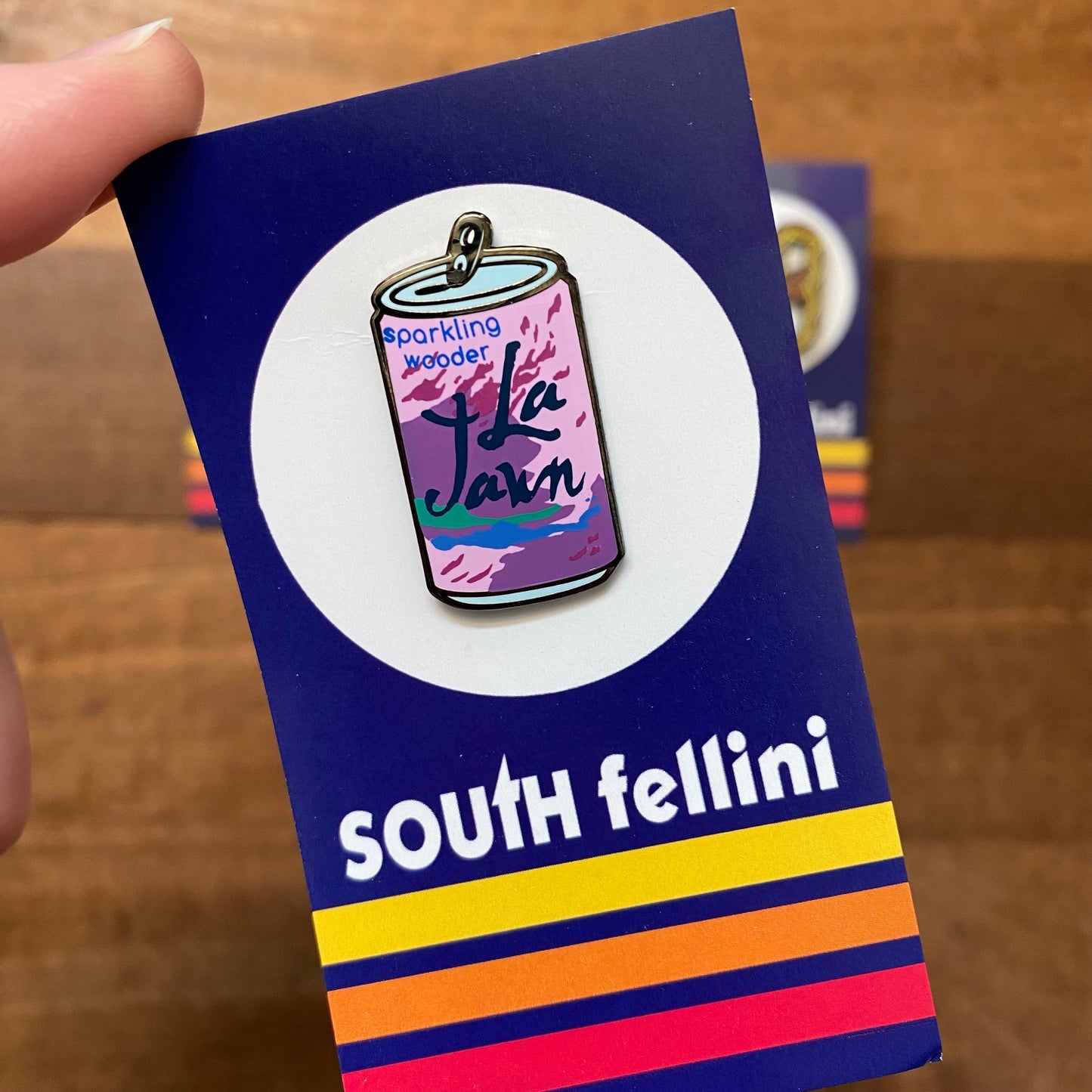 A hand holding a South Fellini Philly-Themed Enamel Pin with a design resembling a can of sparkling water, against a vibrant striped background.