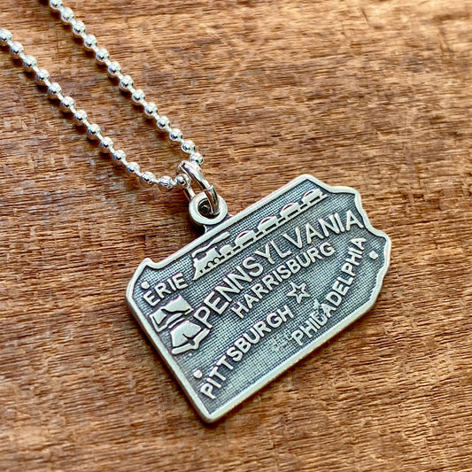 The Pennsylvania Necklace by Stone Cooper features a sterling silver metal pendant shaped like the state of Pennsylvania, showcasing major cities such as Erie, Harrisburg, Pittsburgh, and Philadelphia on a beaded chain. This unique piece offers a timeless look.