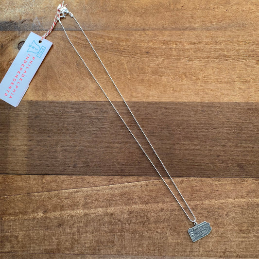 The Pennsylvania Necklace by Stone Cooper, a sterling silver piece featuring a small rectangular pendant reminiscent of the Pennsylvania state shape, is displayed on a brown wooden surface. A pink hang tag reading 