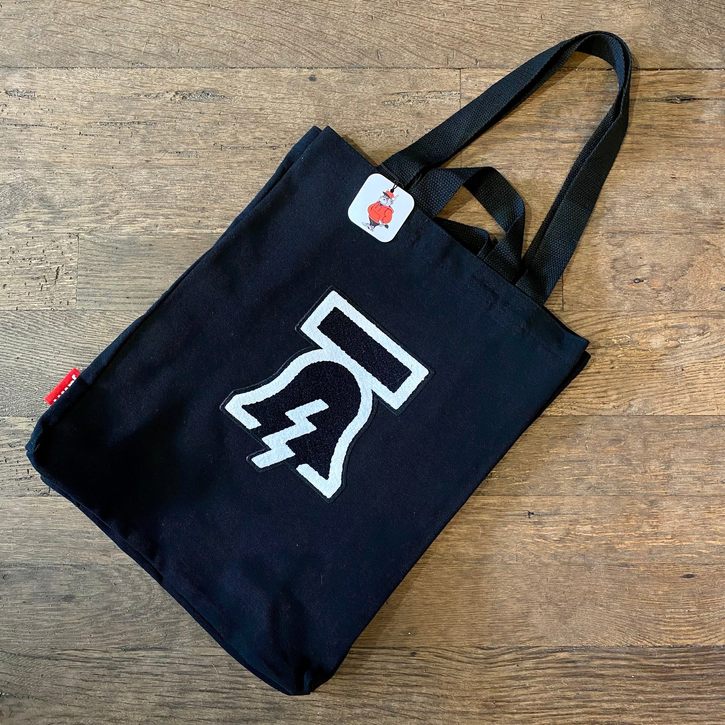 Bell & Bolt Patch Tote Bag by Tote Jawn with white lightning bolt design on wooden floor.
