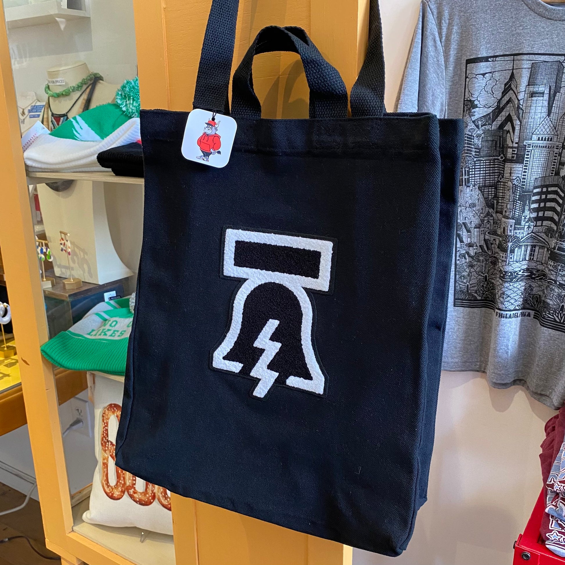 Black Bell & Bolt Patch Tote Bag with a white hourglass and lightning bolt design hanging on a display hook by Tote Jawn.