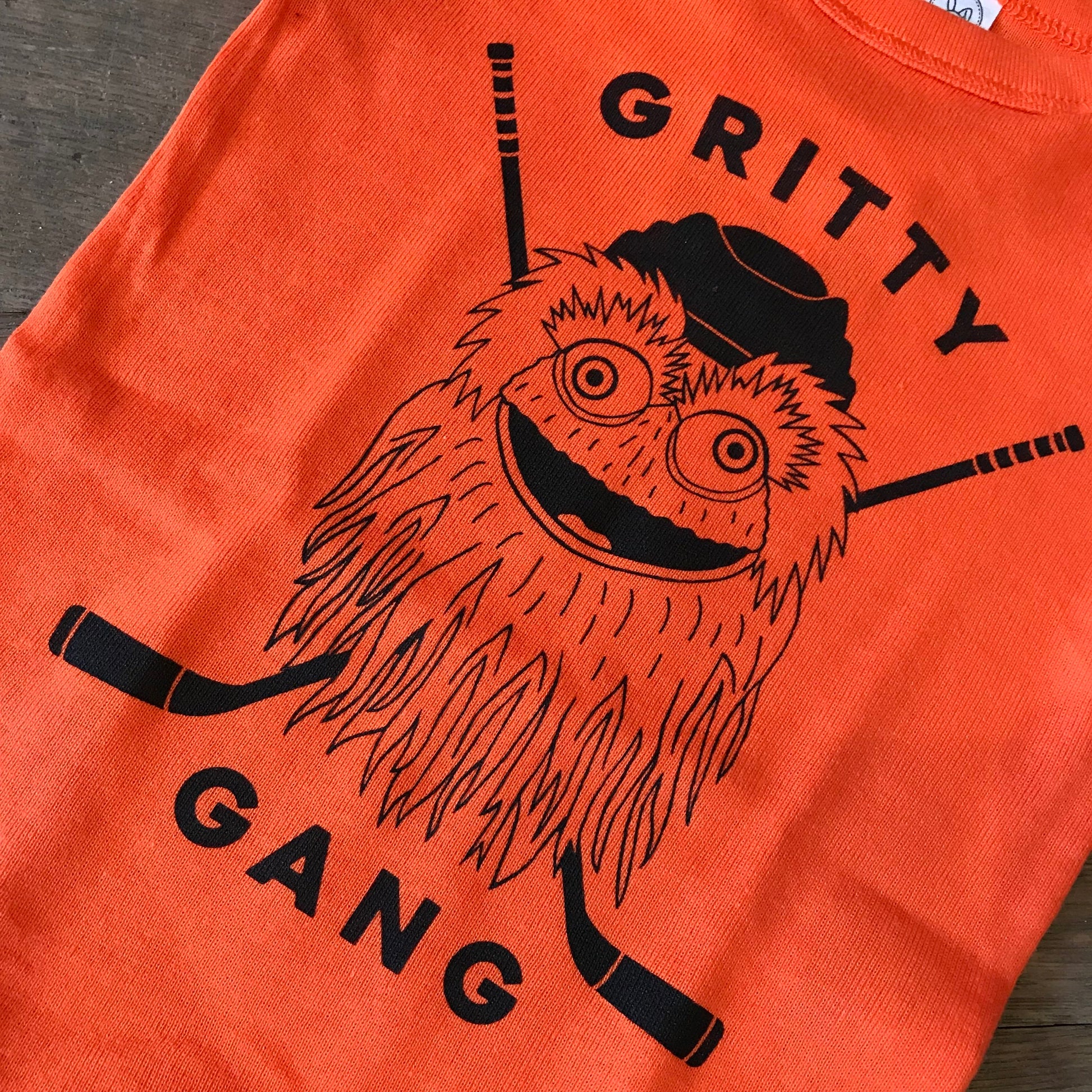 Gritty Gang Baby Onesie featuring an illustration of Philadelphia Flyers mascot GRITTY and the text "gritty gang," with hockey sticks in the background, designed as 100% cotton babywear by exit343design.