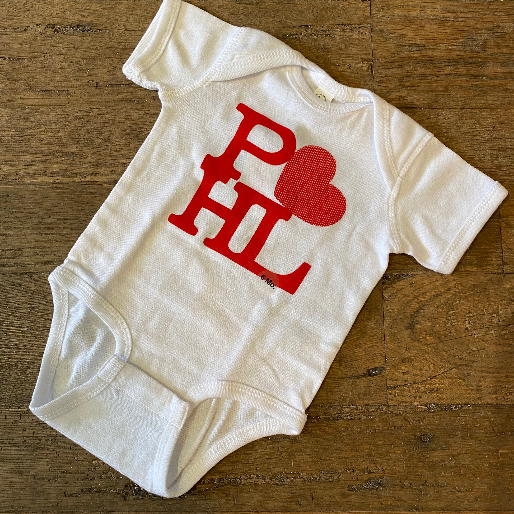 White cotton PHL Love Baby Onesie with red "Philly love" lettering and a heart shape on a wooden surface by exit343design.