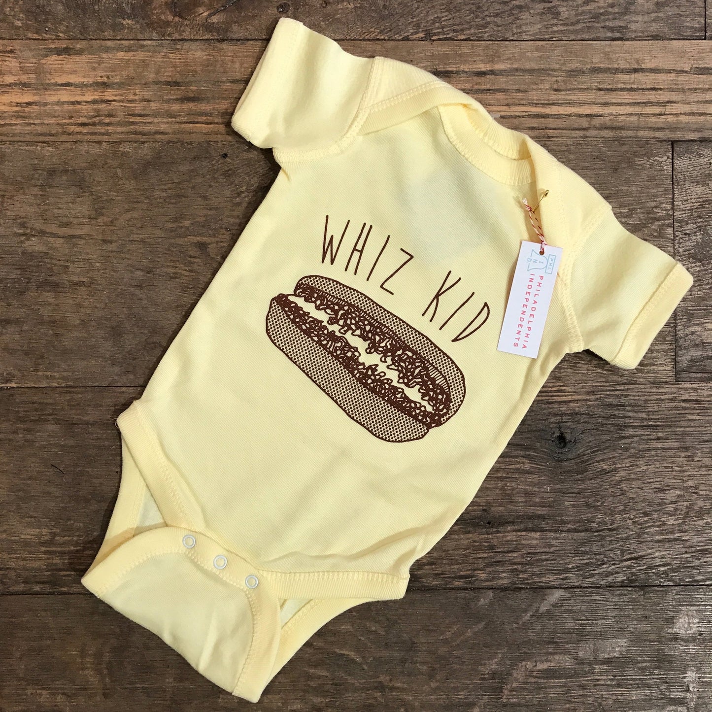 A yellow Whiz Kid Baby Onesie with a cheesesteak-inspired hot dog print and the phrase "Philly genius" displayed on a wooden surface by exit343design.