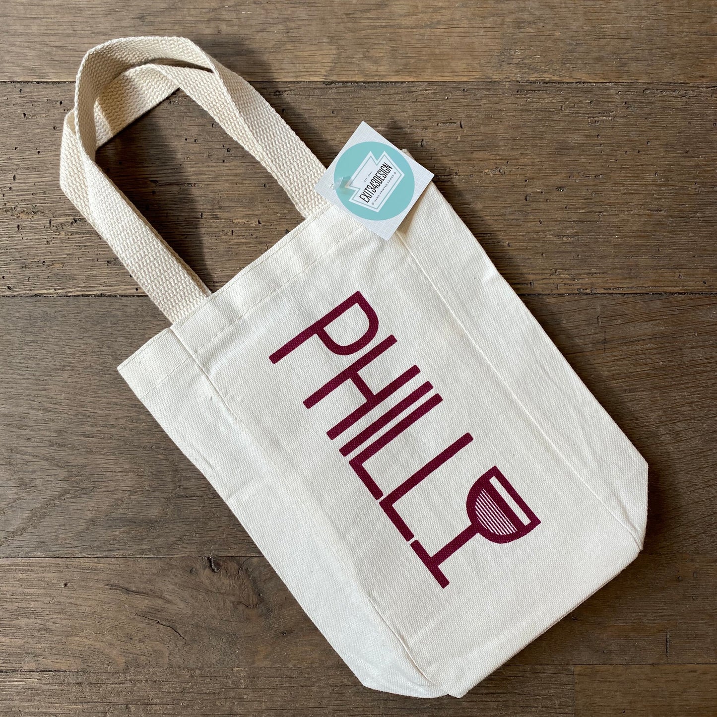 A hand-screenprinted canvas Philly Wine Tote with the word "Philly" printed on it, resting on a wooden floor by exit343design.