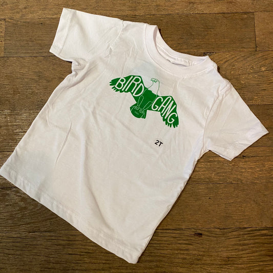 White 100% cotton Bird Gang Toddler T-Shirt with "bird gang" text and bird illustration by exit343design lying on a wooden floor.